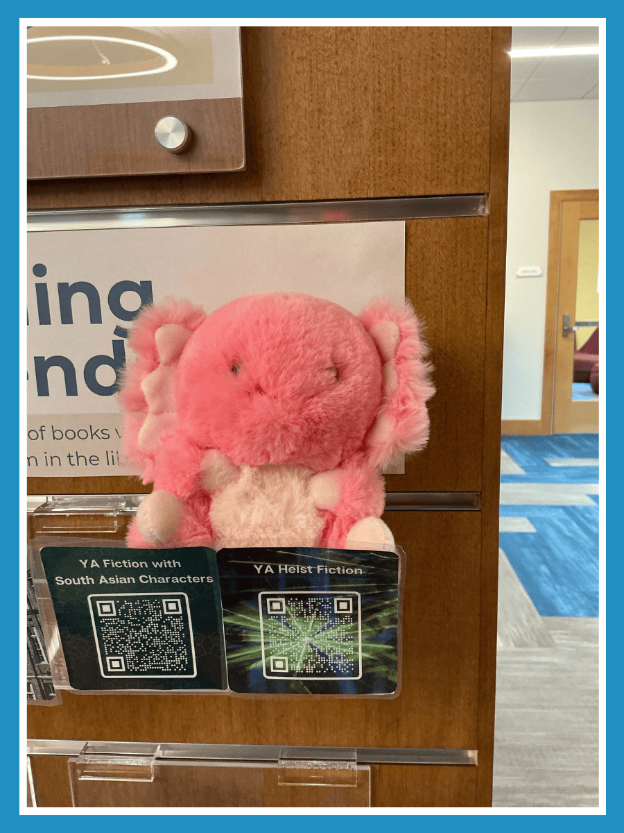 A photo of a stuffed pink axolotl on a bookshelf next to signs with QR codes on them.