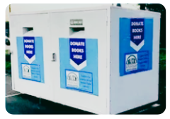 A large white outdoor donation bin with blue signs that read Donate Books Here, placed on a paved surface. The bin has two drop-off slots for book donations.