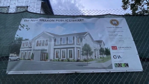 A banner on a chain-link fence displays an architectural rendering of the new Sharon Public Library, showing a two-story building with large windows and landscaping in front. Several sponsor logos are visible on the right side.